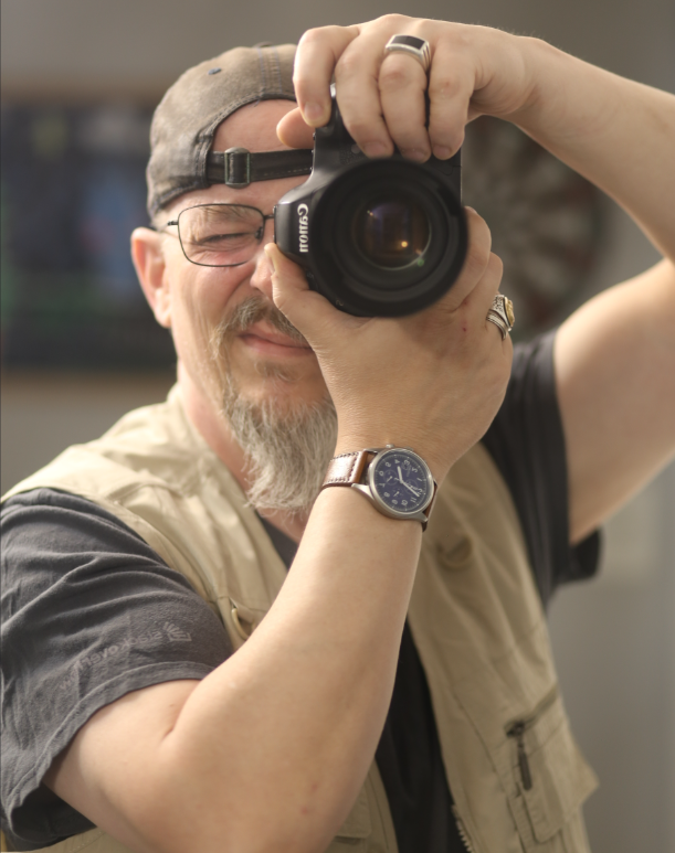 Tim Post taking a photography 'self portrait' by pointing his DSLR at a mirror. He's wearing a field vest with backwards hat while out shooting.